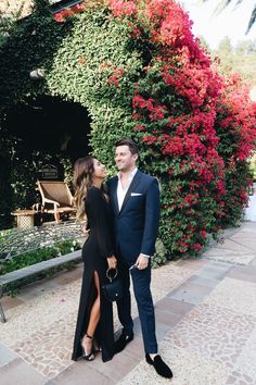 a man and woman standing next to each other in front of some bushes with red flowers