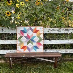 a colorful quilt sitting on top of a wooden bench in front of sunflowers