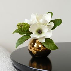 a white flower in a gold vase on a black table next to a white wall