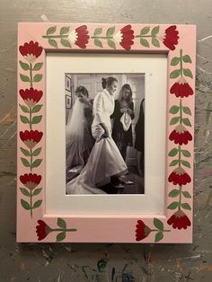 a pink frame with red flowers and leaves on the edge is holding a photo of two women in wedding dresses