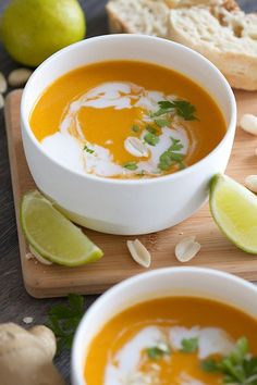 two bowls of carrot soup on a cutting board with limes and bread in the background