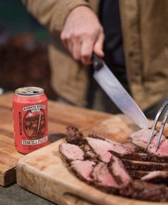 a person cutting meat with a knife on a cutting board next to a can of beer