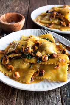 two white plates topped with ravioli covered in cheese and nuts next to a wooden bowl