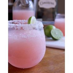 two margaritas sitting on top of a wooden table next to lime wedges and a cutting board