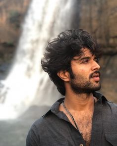 a man standing in front of a waterfall with his hair blowing in the wind and looking off into the distance