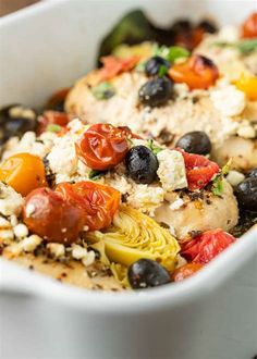 a white dish filled with chicken, olives, tomatoes and artichokes