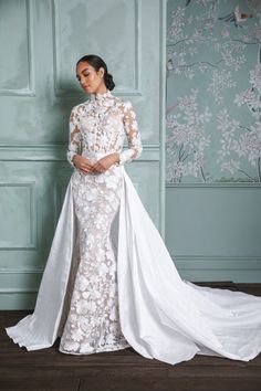 a woman standing in front of a wall wearing a white wedding dress with long sleeves