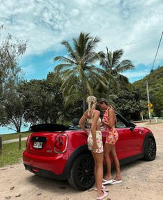 two women standing in front of a red sports car on the side of the road