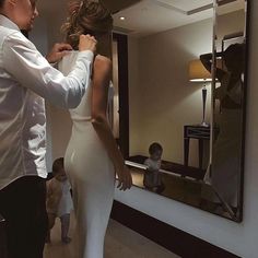 a man helping a woman put on her wedding dress in front of a large mirror