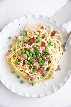 a white plate topped with pasta covered in bacon and green peas next to a fork