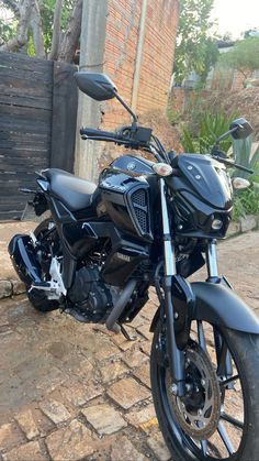 a black motorcycle parked on top of a brick road next to a tree and fence