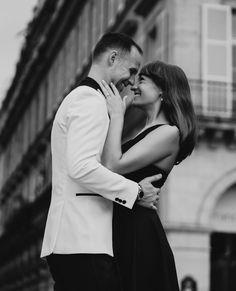 A black and white photo of a couple embracing in front of a building. Couple Embracing, White Artwork, Black And White Artwork