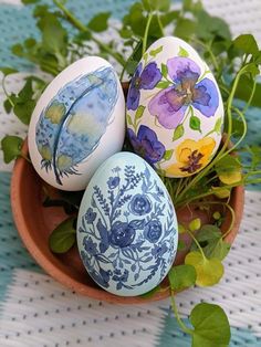 three painted eggs sitting on top of a wooden bowl next to green leaves and flowers