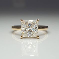 a princess cut diamond sits on a white surface with gold trimmings and an elegant band