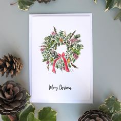 a christmas card with a wreath on it and pine cones next to it, surrounded by greenery