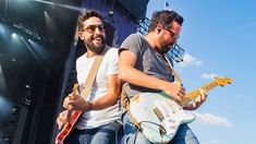 two men are playing guitars on stage at a music festival, one man is wearing sunglasses and the other has a guitar in his hand