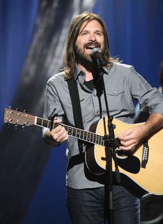 a man holding a guitar while standing in front of a microphone and wearing a gray shirt