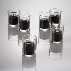 several clear glass vases with black candles in them on a reflective surface, set against a white background