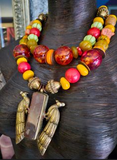 An exotic red, yellow and orange African Statement Necklace is a funky ethnic melange that will enhance your face, beautifully!! This mixed media piece features Tibetan Resin, Brass Chinese Coins, ornate Vintage Brass Beads, Yellow Resin, red magnesite, coral, Picasso Jasper, Nepalese glass, Wood barrels, wavy copper metal spacers, Moroccan Resin, and frosted agates. The 3.5" x 3" Tibetan Brass Repousse Double Peacock Pendant with Inlaid Jasper integrates all the bold and chunky materials and grounds the piece in luxury. Striking worn with a tan or on women if color, year-round. Adjusts from 18-21" with gold tone hardware and a fine hook clasp. Note the funky beaded dangle suspended from the 3" extender chain which will sashay down your back. A high end, dramatic piece from the Atelier of Traditional One-of-a-kind Multicolor Beaded Necklace, Vibrant Orange Jewelry For Festival, Vibrant Orange Festival Jewelry, Multicolor Large Beads Necklace For Rituals, Multicolor Necklaces With Large Beads For Rituals, Artisan Orange Beaded Necklaces For Festivals, Unique Multicolor Decorative Necklaces, Bohemian Orange Necklaces For Festive Occasions, Bohemian Orange Necklace For Festive Occasions