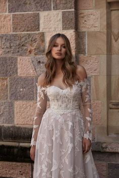 a woman standing in front of a brick wall wearing a white dress with long sleeves