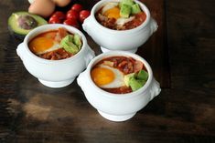 three white bowls filled with chili, eggs and avocado on top of a wooden table