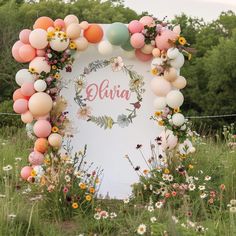 an arch decorated with balloons and flowers in the middle of a field, surrounded by wildflowers