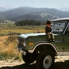 a young boy sitting on the hood of a green pick up truck with mountains in the background