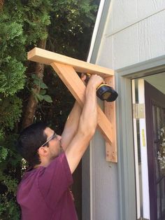 a man working on a wooden cross in front of a house with trees and bushes behind him