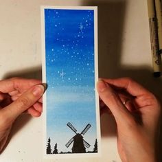 two hands holding up a card with a painting of a windmill in the sky and stars above it