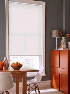 a bowl of fruit sits on a table in front of a large window with white roller shades