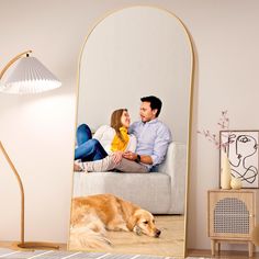 a man and woman sitting on a couch next to a dog in front of a large mirror