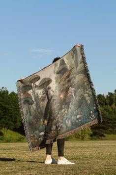 a person standing in the grass with a blanket over their head, holding up a large piece of artwork