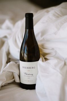 a bottle of wine sitting on top of a white sheet covered bed next to a pillow