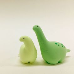 two small plastic dinosaurs sitting next to each other on a white surface, one green and the other light green