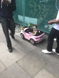 a small child in a pink toy car on the sidewalk with people looking at it