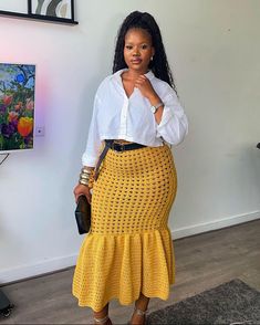 a woman standing in front of a wall wearing a white shirt and yellow crochet skirt