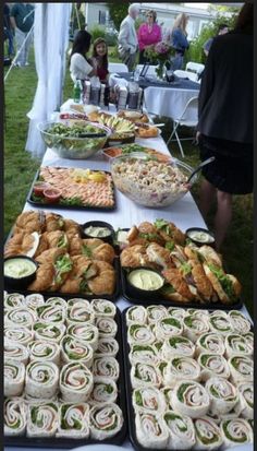 a table full of sandwiches and other food items at an outdoor event with people in the background