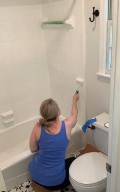 a woman in blue shirt and black pants painting a white wall next to a toilet