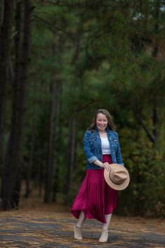 a woman in a red skirt and blue jacket is walking through the woods with a hat on her head