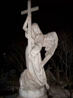 a statue of an angel holding a cross in front of some trees and bushes at night