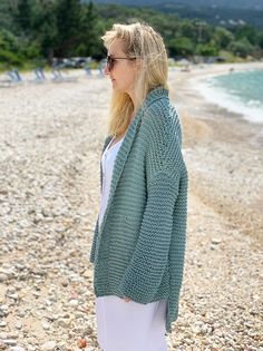 a woman standing on top of a rocky beach next to the ocean wearing a green knitted cardigan