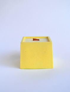 a yellow square candle holder on a white surface with a small wooden stick sticking out of it