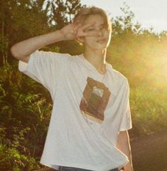 a young man standing in front of trees with his hand on his head and the sun shining behind him