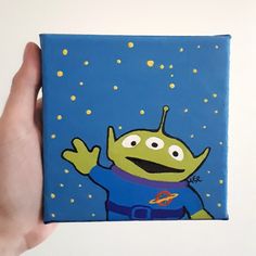 a hand holding up a small square painting with an alien on it