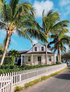 a white house surrounded by palm trees on the side of a road in front of a white picket fence