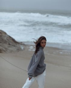 a woman is walking on the beach in white pants and a gray sweater with her hair blowing in the wind