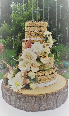 a three tiered cake with flowers and greenery on top is sitting on a tree stump