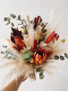 a hand holding a flower arrangement with feathers and flowers on it's side, in front of a white background