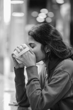 a woman sitting at a table drinking from a paper cup while holding her hands to her face