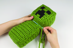 someone is knitting a green knitted bag with a black cross on it and two hands are holding the yarn